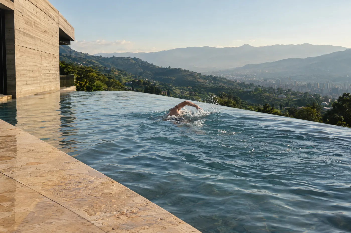 A swimmer in an infinity pool of a modern villa above green mountains in warm morning light.